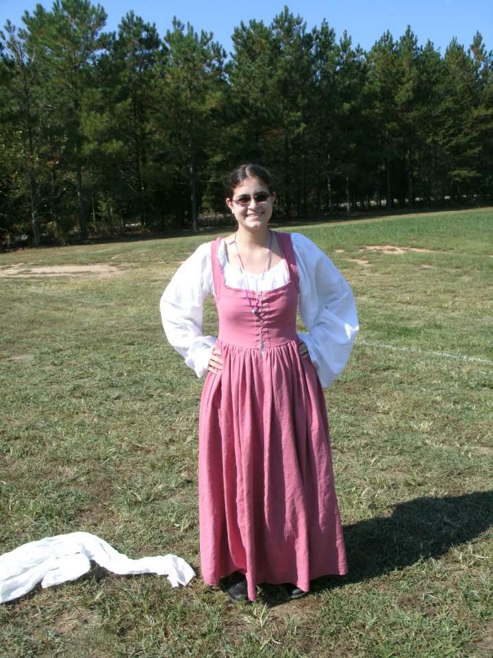 woman standing in a field wearing a white chemise with large sleeves and a sleeveless square-necked pink linen dress, laced up the front